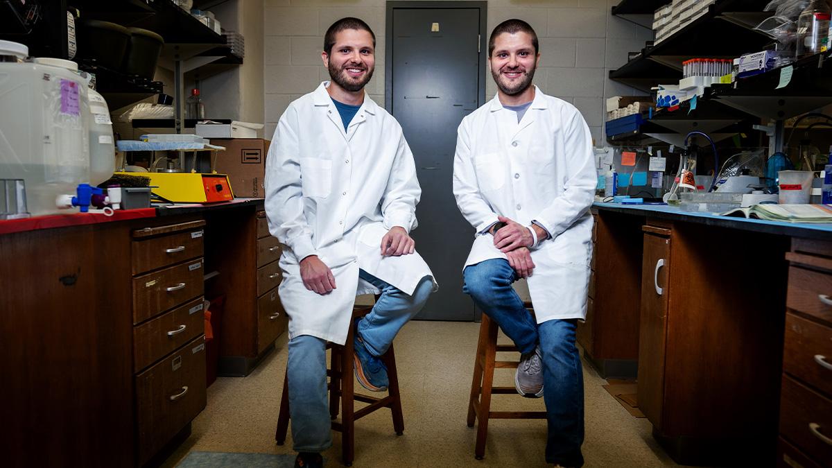 Two twins, Ryan and Brandon Mouery, wearing lab coats and posing for a portrait while sitting on stools in a lab.