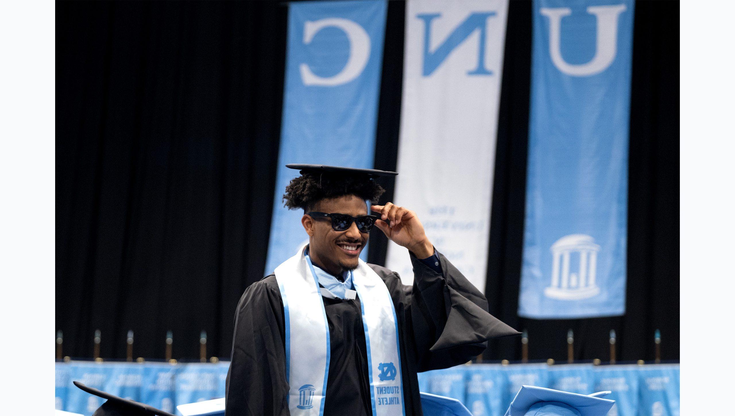 A graduate student in black regalia and wearing sunglasses smiling as he walks at U.N.C. Chapel Hill's Winter Commencement.